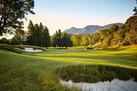 A beautifully maintained golf course with lush green fairways, sand traps, and scattered trees, set against a backdrop of rolling hills and a clear sky at dawn or dusk.