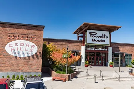 The exterior of a Powell's Books store at Cedar Hills Crossing on a sunny day, featuring a large sign, brick buildings, and some greenery.