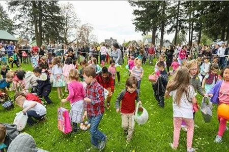 A large group of children excitedly search for Easter eggs scattered across a wide green lawn in a park filled with trees and adults watching in the background.