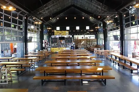 The spacious interior of a food cart station featuring numerous long wooden picnic tables and benches arranged under a high, dark ceiling with string lights. Large garage doors line the walls, allowing natural light to flood the area, and various food vendors are visible in the background.