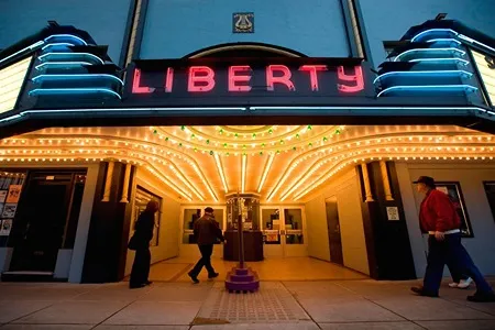 The brightly lit marquee of the Liberty Theatre at dusk, featuring red neon lettering spelling "LIBERTY" and warm white lights illuminating the entrance. People are seen walking into and past the theater.