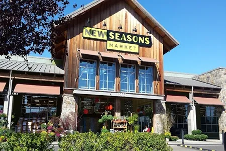 The exterior of a New Seasons Market store on a sunny day, featuring a rustic wooden facade with large windows displaying produce and floral arrangements. A bright "NEW SEASONS MARKET" sign is visible above the entrance.