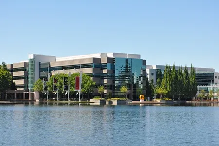 A sprawling complex of modern office buildings with glass and metal facades, situated along the edge of a calm blue lake under a clear sky. Lush green trees surround the buildings and the waterfront.