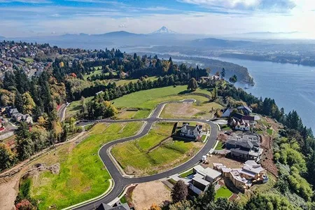 An aerial view of a residential area situated on rolling green hills overlooking a wide blue river and distant mountain ranges under a partly cloudy sky. Some houses are built, and others are under construction along winding roads.